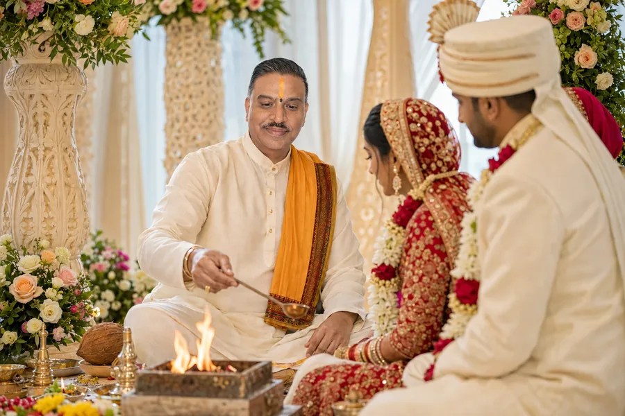 A priest making an offering into the sacred fire as a couple sit beside him beneath a floral mandap