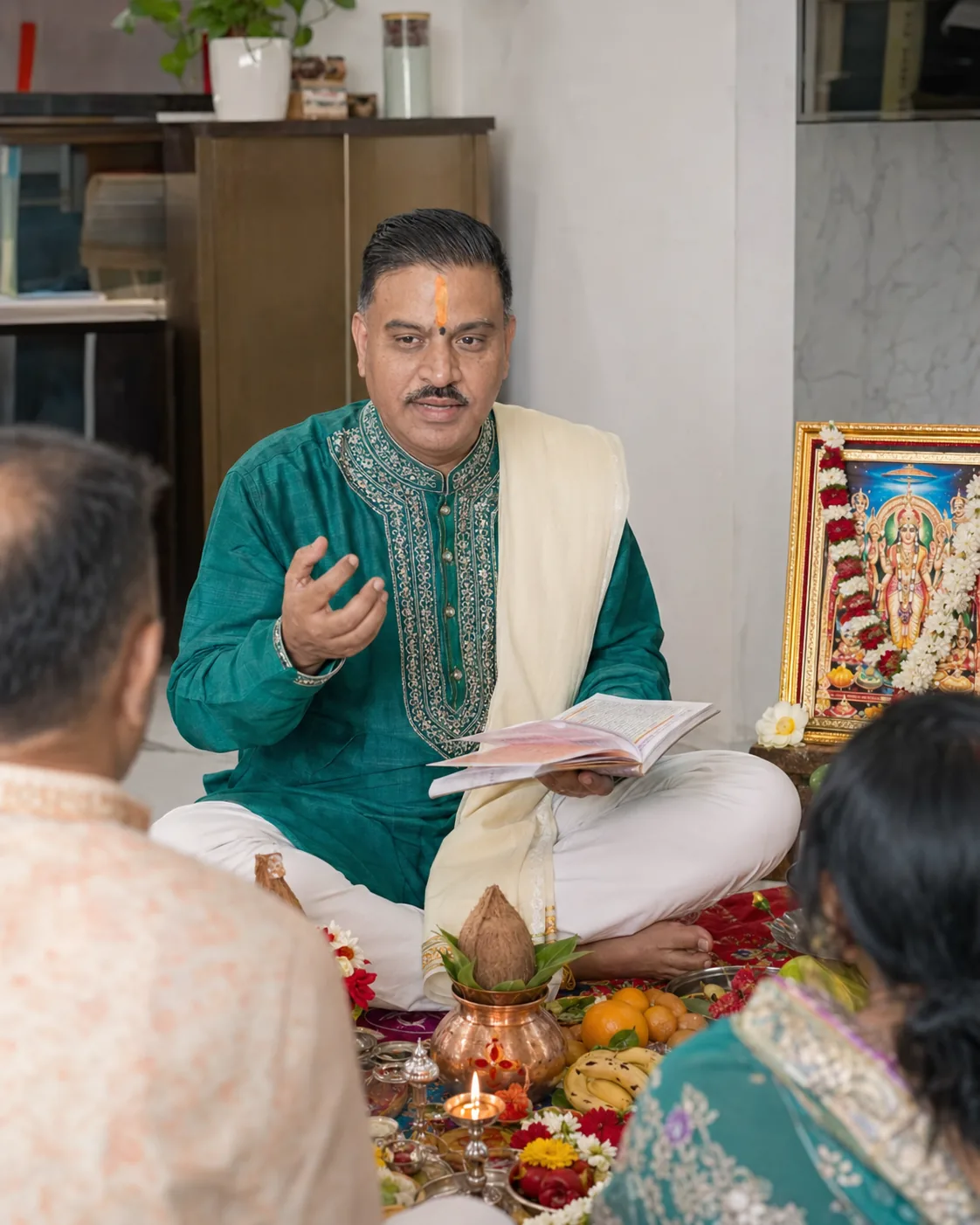 A Hindu priest seated with an open ritual text beside a framed image of Lord Vishnu, a kalash, a lit diya, flowers, fruit and puja materials, with family members gathered in the foreground